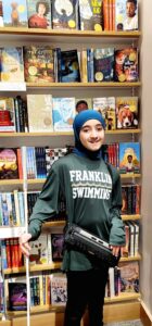 The individual is wearing a teal hijab, a dark green long-sleeved sweatshirt with 