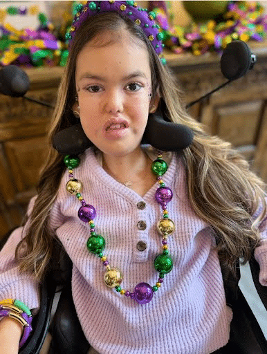 Picture of girl in wheelchair wearing Mardis Gras Beads and a purple shirt.
